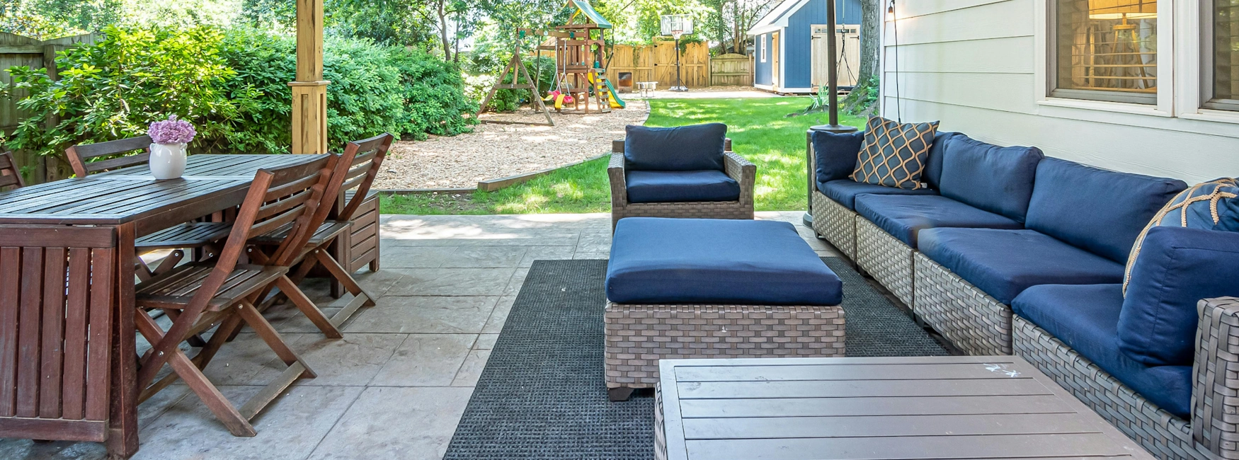 Backyard patio in a Buckmaster’s Circle home with wicker sofa and navy cushions, wooden dining set, and a play-set visible in the grassy yard beyond