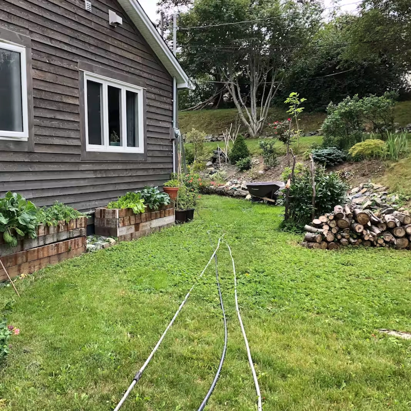 Backyard in Churchill Park with raised garden beds, firewood stack, a wheelbarrow, and a sloped, landscaped hill.