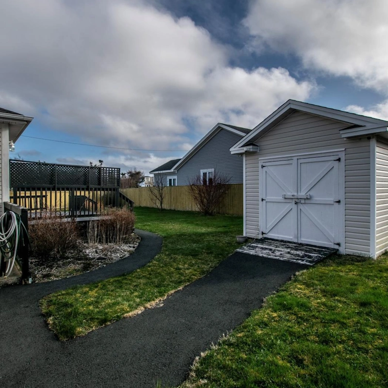 Backyard of a Clovelly Trails home for sale, featuring a storage shed, raised deck, paved walkway, and fenced lawn under a cloudy sky.
